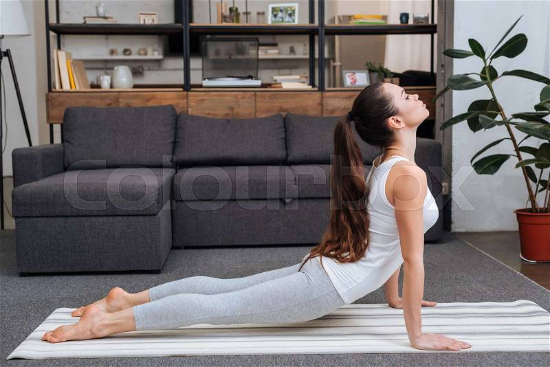 Young woman practicing cobra pose in living room at home, stock photo