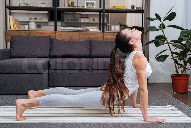 Young woman practicing cobra pose in living room at home, stock photo