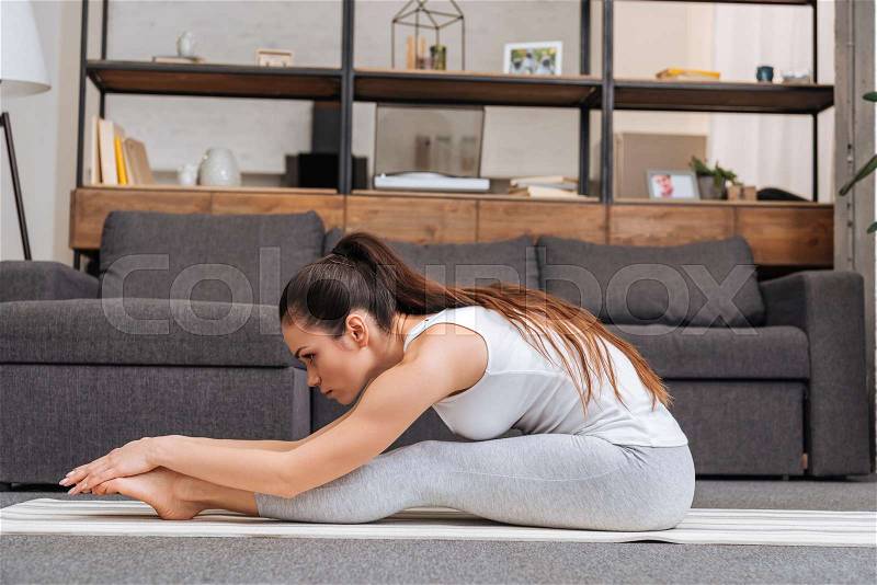 Woman practicing seated forward fold pose on fitness mat at home in living room, stock photo