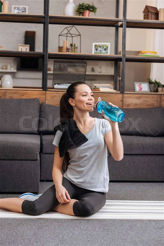Sportswoman sitting on fitness mat and drinking water from sport bottle in living room , stock photo