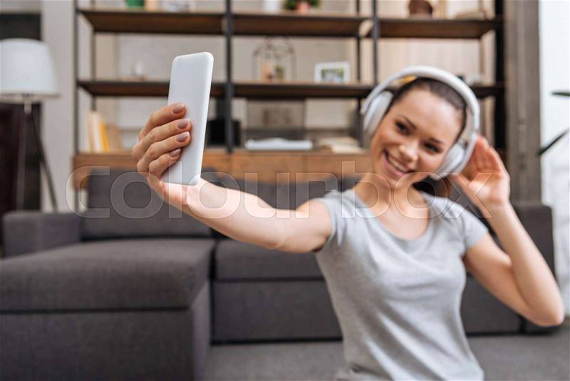 Beautiful smiling woman in headphones taking selfie on smartphone at home, stock photo