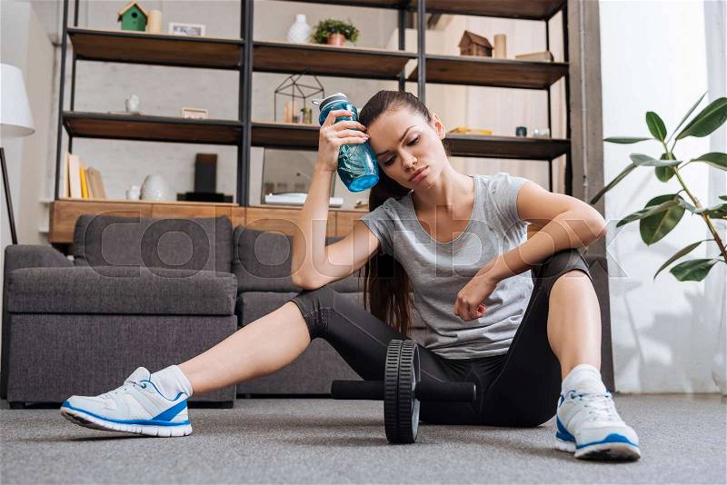 Tired sportswoman with sport bottle after training with ab wheel at home, stock photo