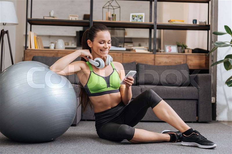Smiling sportswoman in headphones sitting with fitness ball and using smartphone at home, stock photo