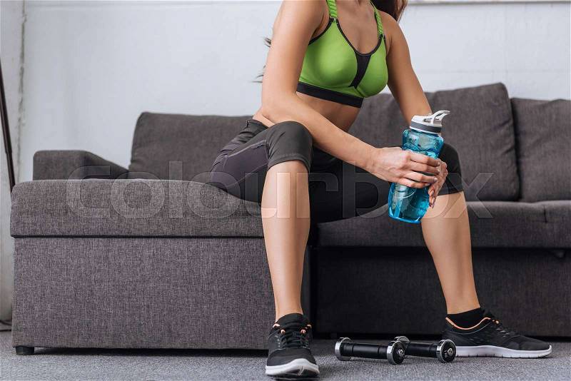 Partial view of sportswoman with sport bottle sitting on couch at home, stock photo
