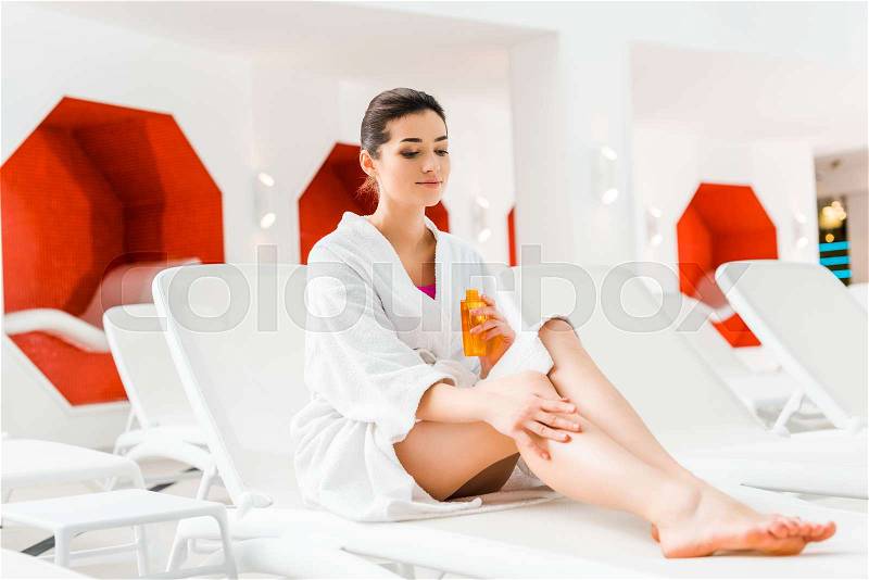 Beautiful woman applying body oil on leg while holding bottle in hand , stock photo