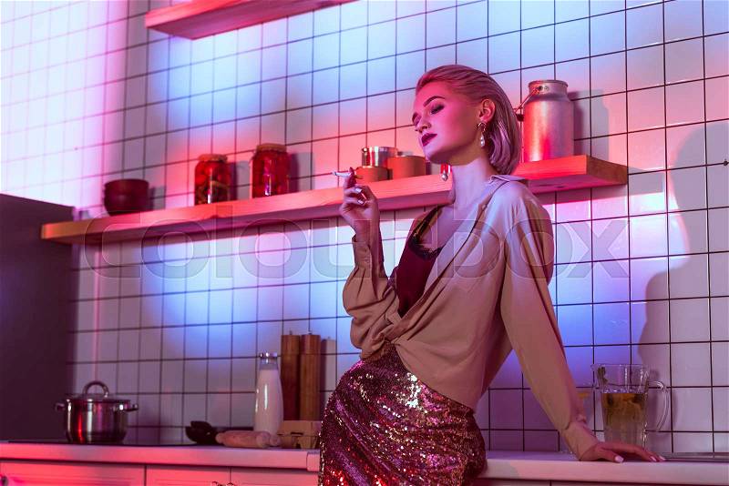 Beautiful fashionable woman smoking in neon light in kitchen with copy space, stock photo