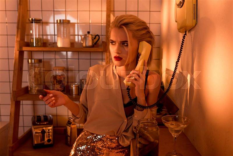Beautiful confused woman talking on retro telephone in kitchen with orange light, stock photo