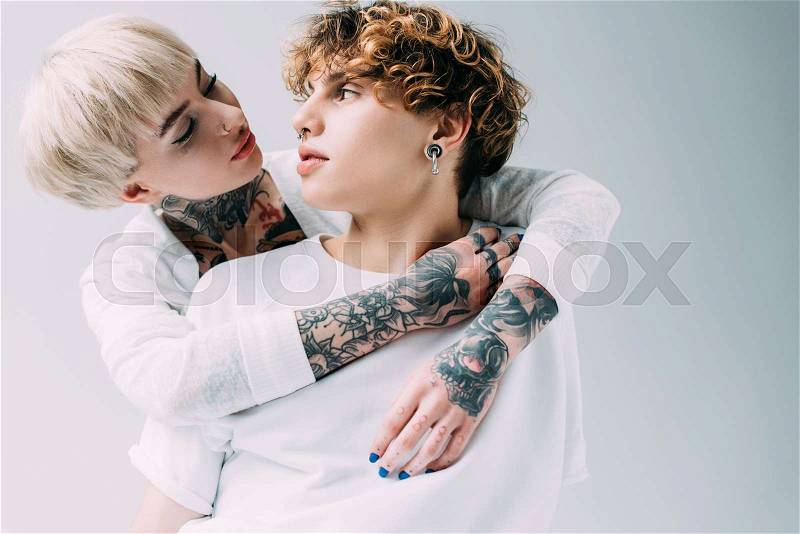 Blonde girl with tattoos looking at boyfriend with curly hair isolated on grey, stock photo