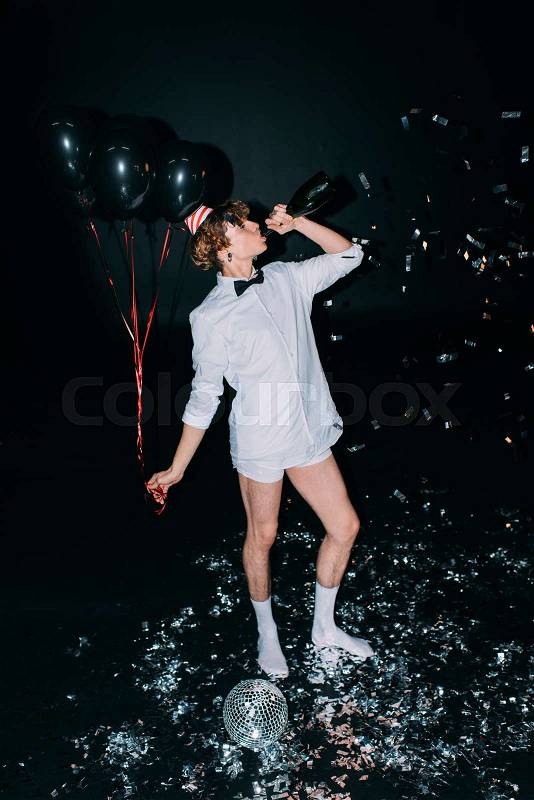 Man with curly hair drinking champagne from bottle and holding balloons isolated on black, stock photo