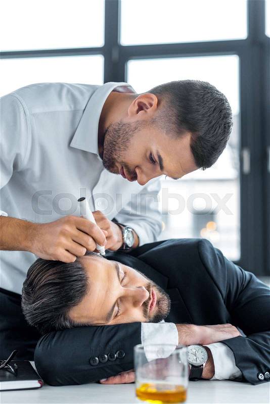 Man drawing with marker on face of sleeping coworker in modern office , stock photo
