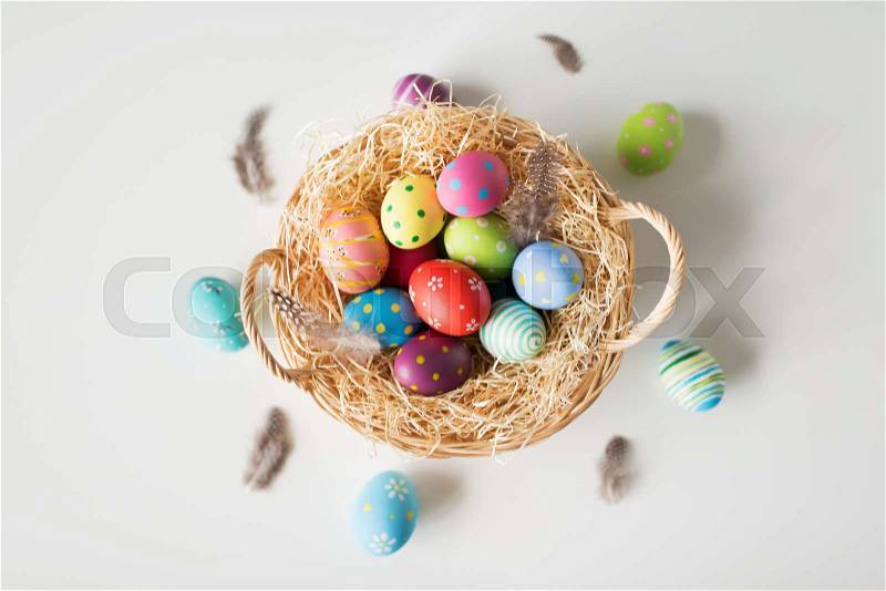 Easter, holidays and tradition concept - colored eggs in basket with straw and quail feathers on white background, stock photo