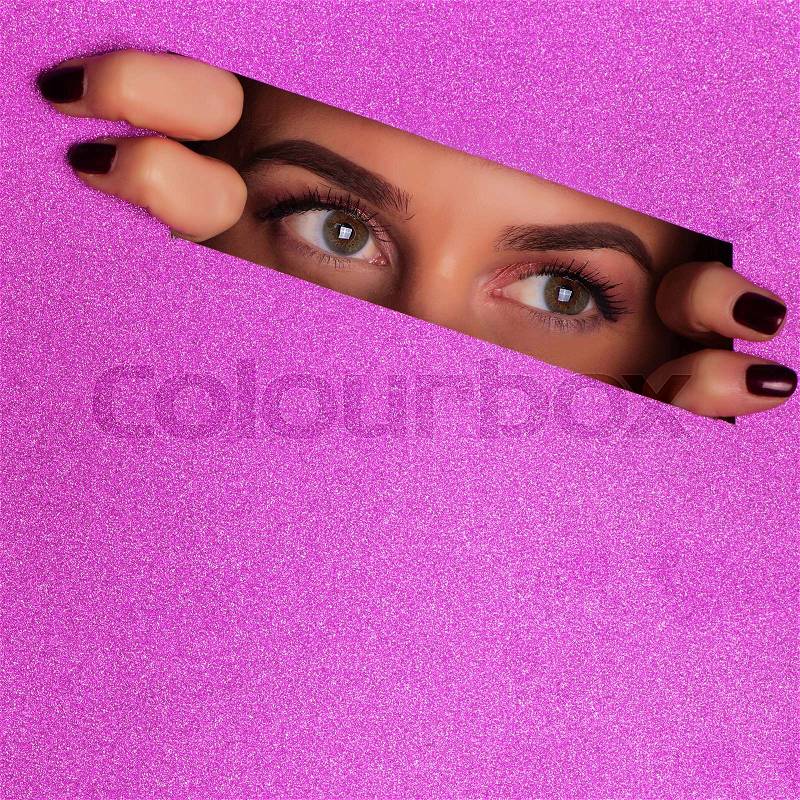 Girl with bright make up looking through hole in violet paper background. Spying, glancing concept. Make up artist, beauty trend. Square crop. Beauty salon ..., stock photo
