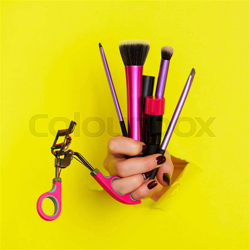 Woman hand with professional cosmetic tools for make up: brushes, mascara, lipstick, eyelash curler on yellow background. Beauty concept for cosmetics sale. Square ..., stock photo