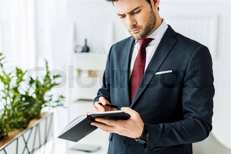 Handsome businessman in formal wear writing in notebook in office, stock photo