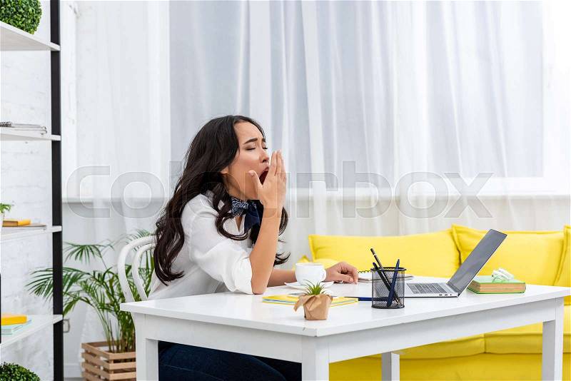 Sleepy asian freelance sitting at work desk and yawing with covering mouth with hand, stock photo