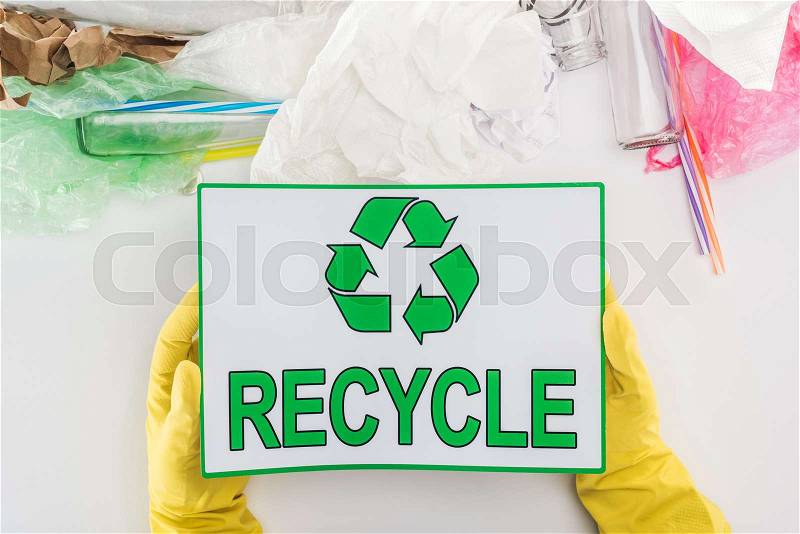 Partial view of man in yellow rubber gloves holding recycling sign among glass bottles, plastic bags, paper and plastic tubes , stock photo