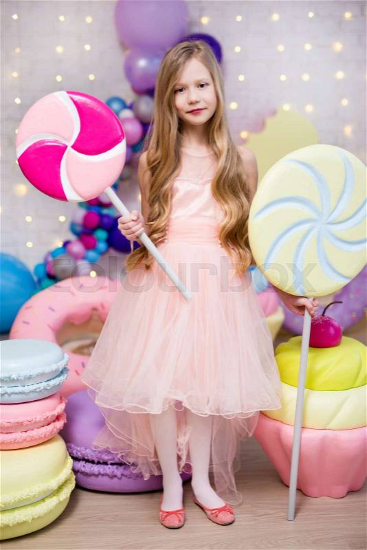 Portrait of cute little girl holding huge lollipops with giant donuts and cupcakes decorations, stock photo