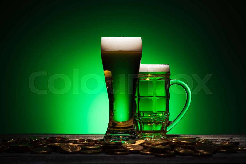 Glasses of irish beer standing near golden coins on wooden table on green background, stock photo