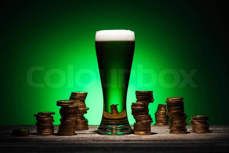 Glass of irish ale standing on wooden table near golden coins on green background, stock photo
