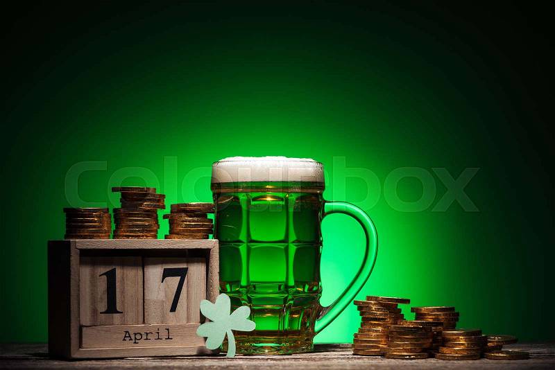 Glass of green irish beer near golden coins and cube calendar on green background, stock photo