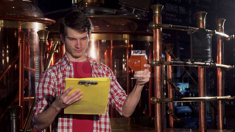 Young man with clipboard working at craft brewery, examining beer. Attractive cheerful male brewer enjoying working at his small brewery. Entrepreneurship, craft ..., stock photo