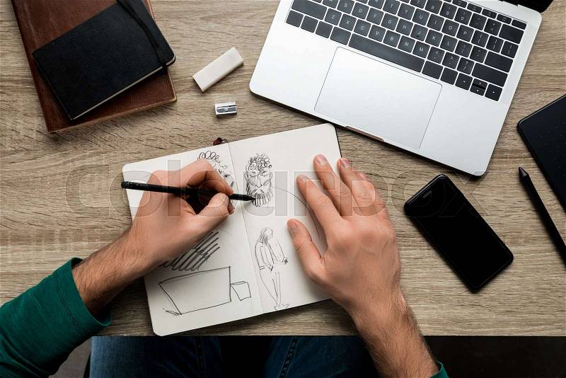Top view of mans hands drawing on album and smartphone next to laptop on wooden table, stock photo
