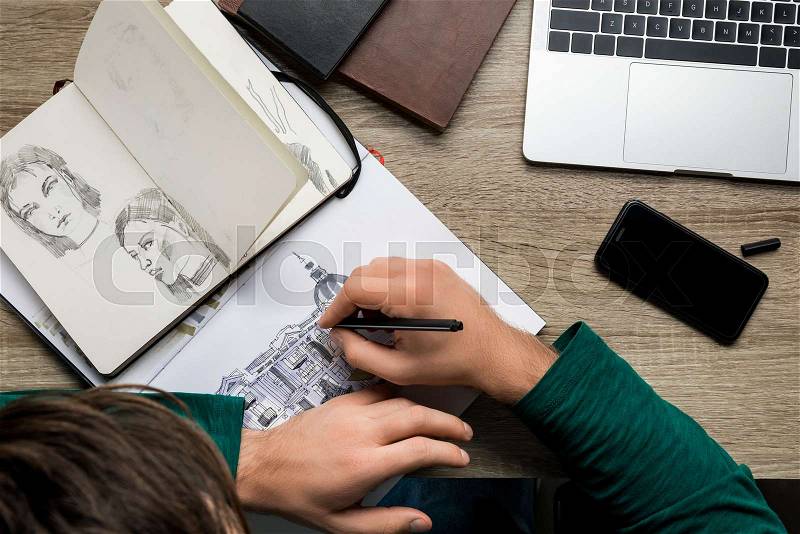 Overhead back view of man drawing in album on wooden table next to laptop and smartphone, stock photo