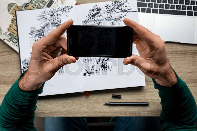 Partial view of man taking drawing photo on smartphone from album lying on wooden table, stock photo