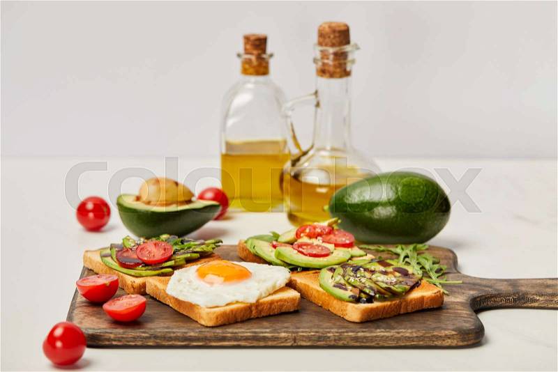 Selective focus of wooden cutting board with toasts, scrambled egg, cherry tomatoes, avocados and oil bottles on grey background, stock photo