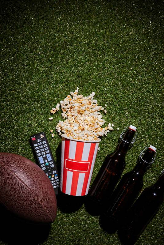 Top view of bottles of beer near popcorn, remote control and ball lying on grass, stock photo