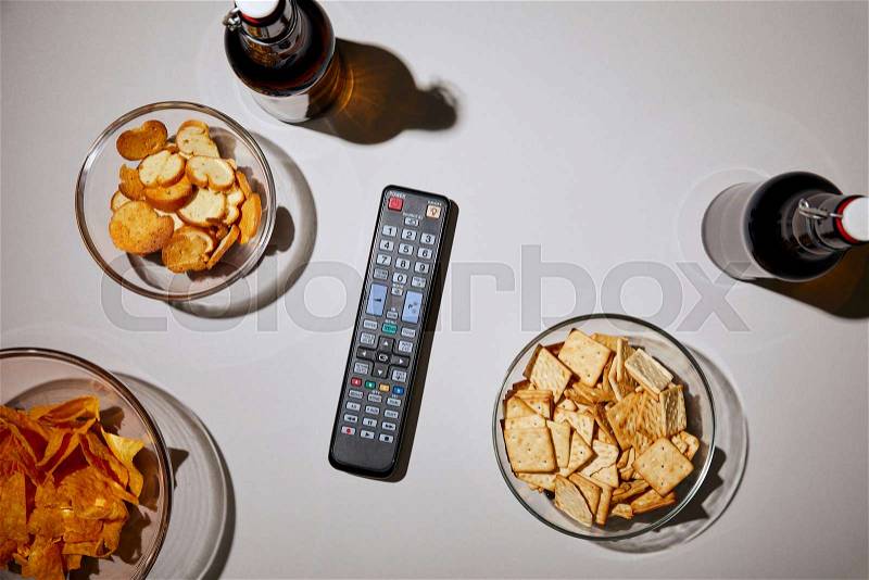Top view of bottles with beer near remote control and snacks on white background, stock photo