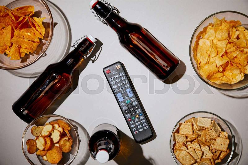Top view of brown bottles with beer near snacks in bowls and remote control on white background, stock photo