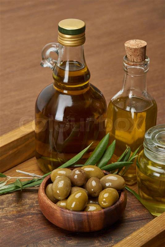 Wooden tray with different oil bottles, bowl of olives and olive tree leaves on brown surface , stock photo