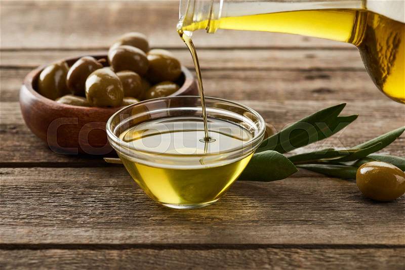 Pouring oil from bottle into glass bowl, bowl of olives, olive tree branch and olive on wooden surface, stock photo