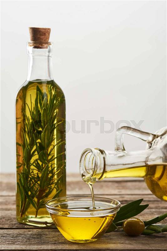 Pouring olive oil from bottle into glass bowl, bottle of oil flavored with rosemary, olive tree leaves and olives on wooden surface, stock photo