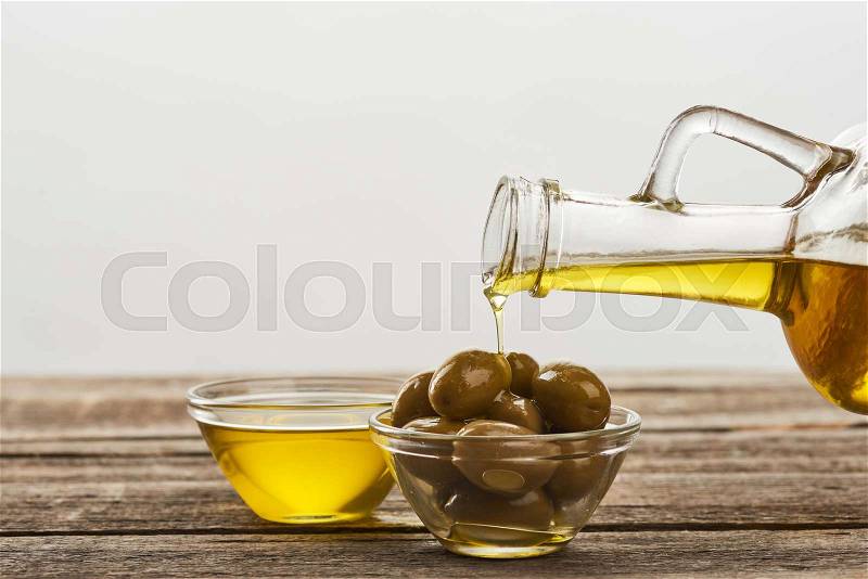 Glass bowl full oil, and pouring of oil into glass bowl with olives on wooden surface, stock photo