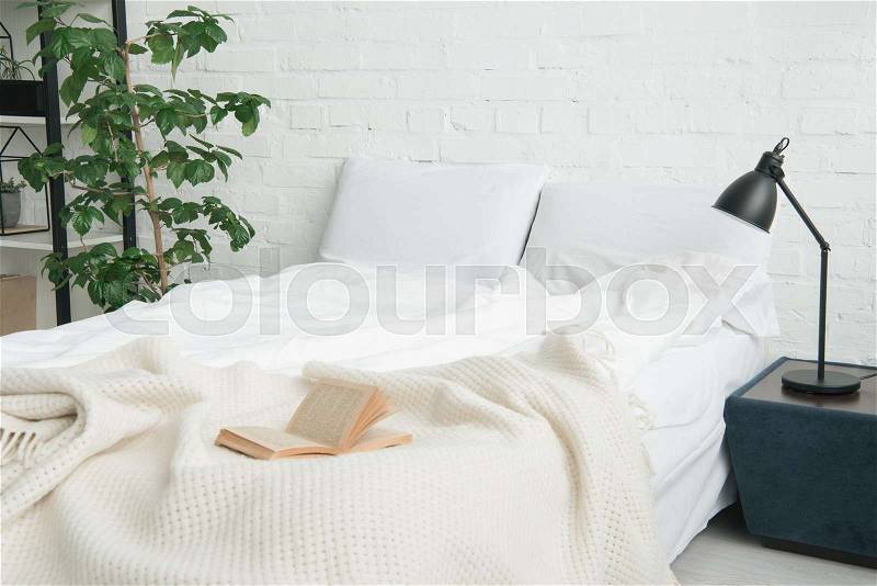 Book on white bed, plant and lamp on black nightstand, stock photo