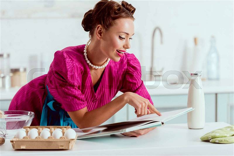 Elegant smiling pin up girl reading recipes book while leaning on kitchen table , stock photo