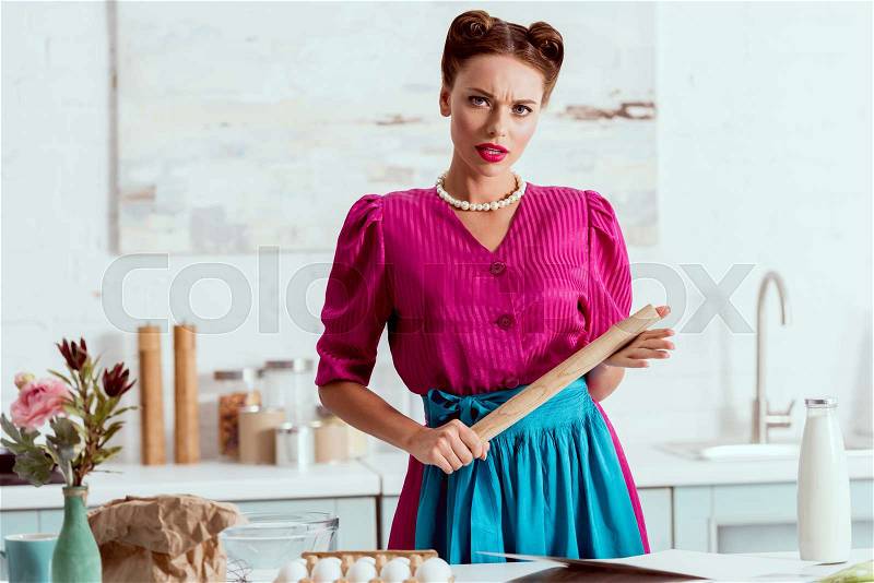 Serious pin up girl holding wooden rolling pin while standing by kitchen table, stock photo