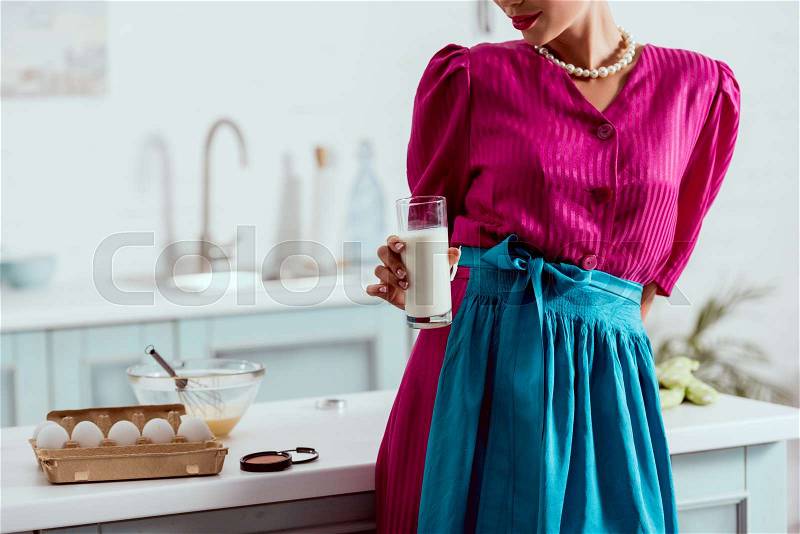 Partial view of elegant pin up girl holding glass of milk while standing near kitchen table with different products and face powder, stock photo