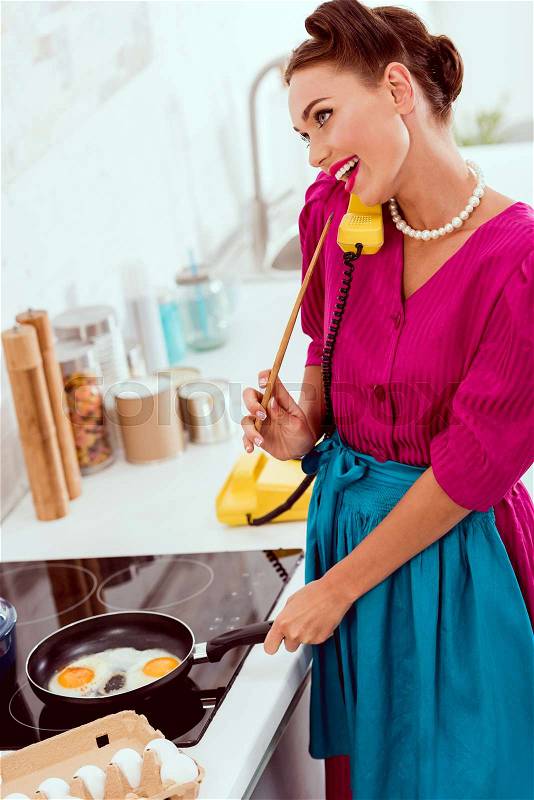 Pretty pin up girl talking on vintage yellow phone while frying eggs, stock photo