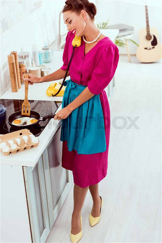 Smiling pin up girl talkin on vintage yellow phone while frying eggs, stock photo