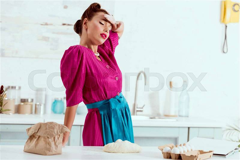 Exhausted pin up girl with flour traces on hands and face resting by kitchen table , stock photo