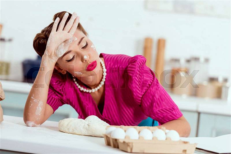 Exhausted pin up girl with flour traces on hands and face leaning on kitchen table, stock photo