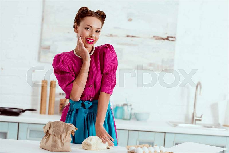 Pretty pin up girl kneading dough on kitchen table and touching face with flour traces, stock photo