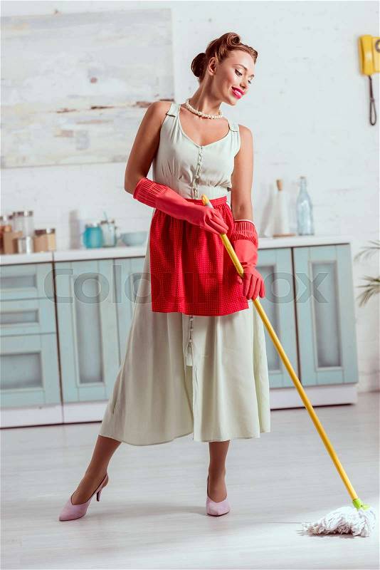Smiling pin up cleaning kitchen floor with yellow mop, stock photo
