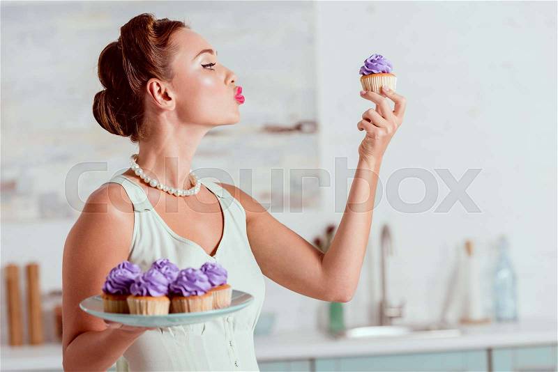 Beautiful pin up girl holding plate full of cupcakes and one cupcake in front of face, stock photo