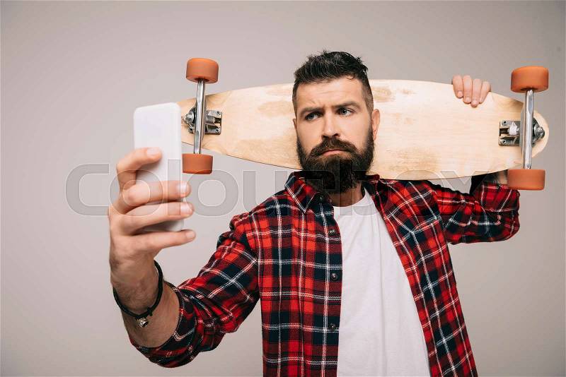 Handsome bearded man in checkered shirt holding longboard and taking selfie on smartphone isolated on grey, stock photo