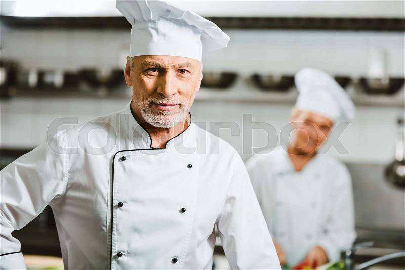 Handsome male chef in uniform and cap looking at camera in restaurant kitchen with copy space, stock photo