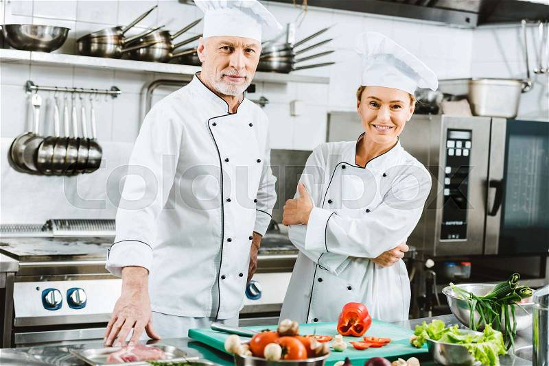 Female and male chefs in uniform looking at camera during cooking in restaurant kitchen, stock photo